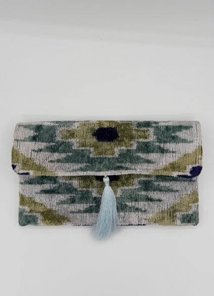 A small woven clutch or wallet with a geometric ikat pattern in olive green, navy blue, and cream, featuring a light blue tassel closure, shown against a plain white background.