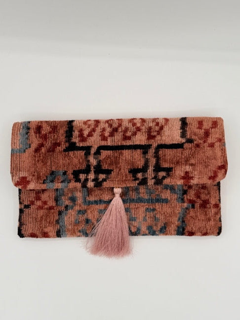 A folded vintage-style patterned rug with a dusty pink tassel accent, displayed against a plain off-white background.