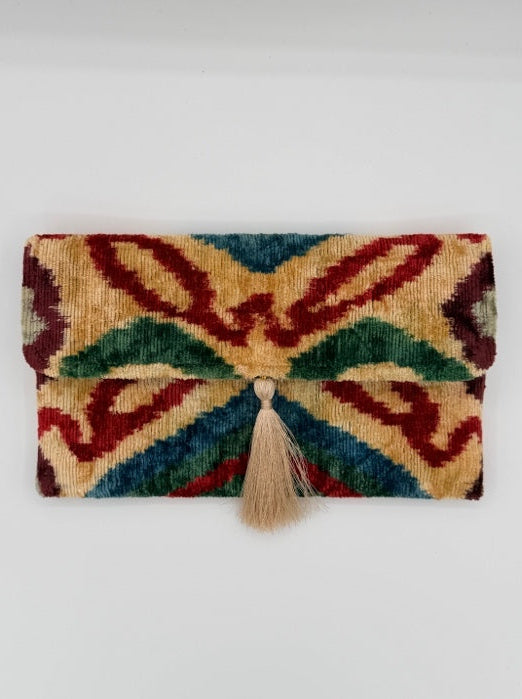 A folded woven textile with an abstract ikat-style pattern in red, green, blue, and beige, secured with a small beige tassel on a plain white background.
