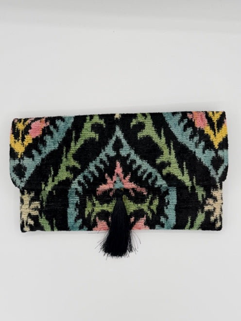 A black woven fabric pouch or clutch with a colorful geometric ikat pattern in pink, green, yellow, and blue, featuring a black tassel closure on the right side, shown against a plain white background.