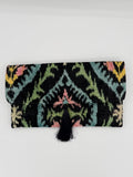 A black woven fabric pouch or clutch with a colorful geometric ikat pattern in pink, green, yellow, and blue, featuring a black tassel closure on the right side, shown against a plain white background.