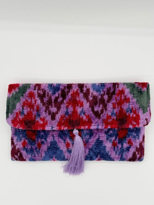 A folded clutch likely a clutch or notebook cover, featuring a vibrant multicolor pattern of red, purple, blue, and green. It has a light purple tassel closure on the right side and is photographed against a plain white background.