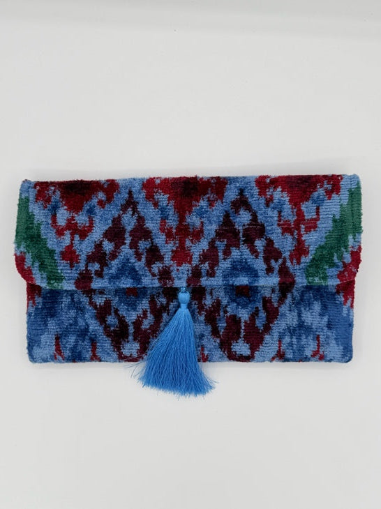 A folded multicolor knit blanket or scarf with a geometric pattern in blue, red, and green, featuring a blue tassel tie on the right side, set against a plain light gray background.