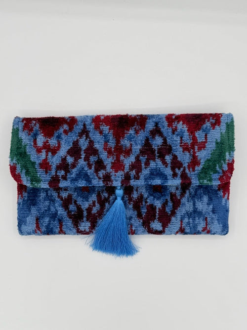 A folded multicolor knit blanket or scarf with a geometric pattern in blue, red, and green, featuring a blue tassel tie on the right side, set against a plain light gray background.