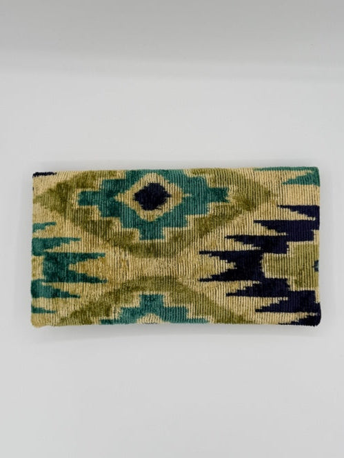 A rectangular woven textile with an ikat-style geometric pattern in olive green, teal, dark navy, and beige, shown hanging against a plain light gray wall.