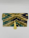 A folded woven textile with an ikat-style geometric pattern in olive green, teal, and dark navy, finished with a light yellow tassel tie, shown against a plain light gray background.