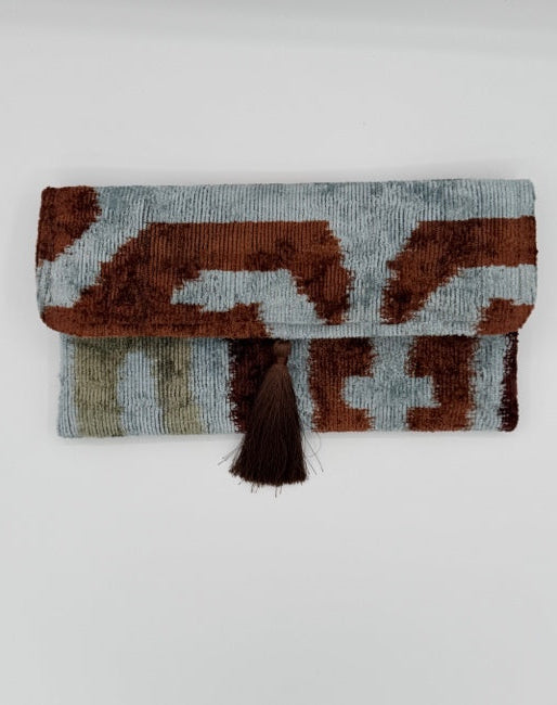 A folded woven textile with an abstract ikat-style pattern in brown, light blue-gray, and beige, featuring a dark brown tassel on the side, photographed against a plain light gray background.