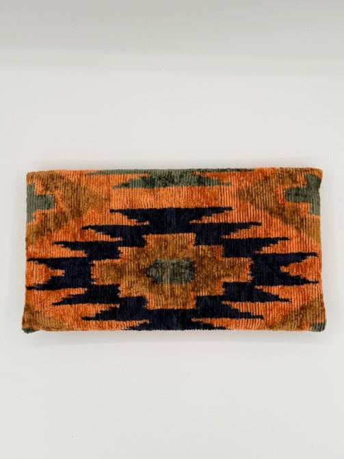 A rectangular woven textile with a geometric ikat-style pattern in orange, black, and muted olive green, shown hanging against a plain white wall.