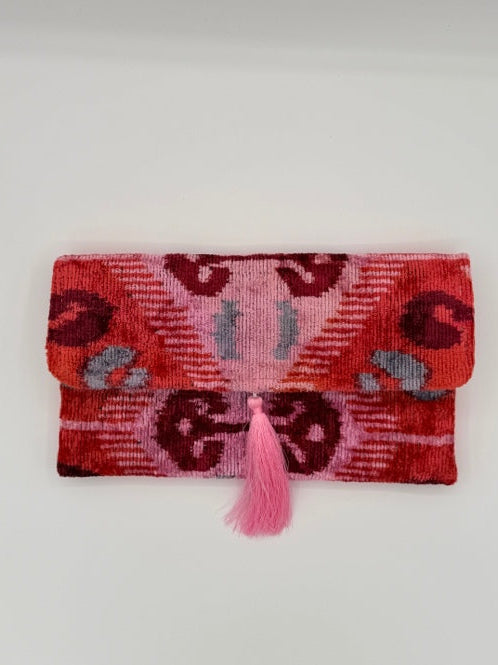 A folded woven textile clutch with a pink tassel, featuring a red, pink, and dark maroon ikat-style pattern on a plain white background.