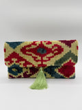 A folded woven textile clutch with an ornate geometric pattern in red, green, blue, and beige, finished with a light green tassel on a plain light gray background.