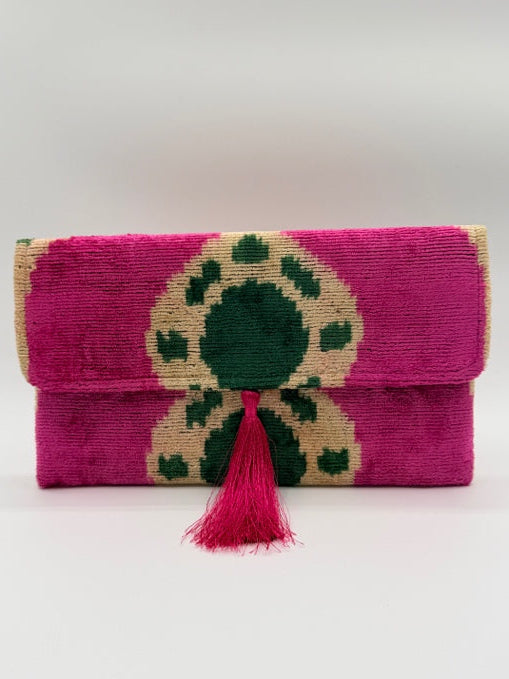 A folded handwoven textile pouch or clutch in vibrant magenta pink, featuring a large central geometric pattern in dark green and natural beige, finished with a matching pink tassel tie on a plain white background.
