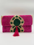 A folded handwoven textile pouch or clutch in vibrant magenta pink, featuring a large central geometric pattern in dark green and natural beige, finished with a matching pink tassel tie on a plain white background.
