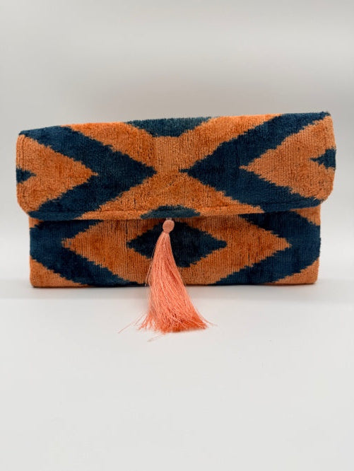 A folded woven textile, likely a towel or blanket, featuring a bold geometric ikat pattern in orange and dark navy blue, finished with a matching orange tassel.