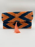 A folded woven textile, likely a towel or blanket, featuring a bold geometric ikat pattern in orange and dark navy blue, finished with a matching orange tassel.