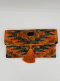 A folded clutch purse with a textured, woven ikat-style pattern in orange, dark green, and brown, featuring a matching orange tassel tie closure, shown against a plain white background.